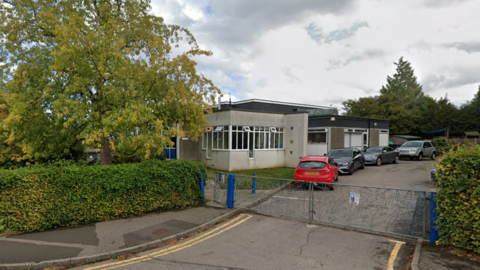 A grey building behind blue gates. It is off a leafy road and cars and parked outside. 