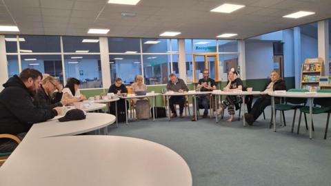 A group of men and women sitting at tables placed in a horseshoe arrangement, engaged in a meeting. All have mugs in front of them. The space has a grey blue carpet and an internal window behind into an area marked reception.