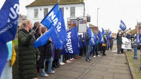 Dozens of people in coats line the street waving blue NASUWT branded flags.