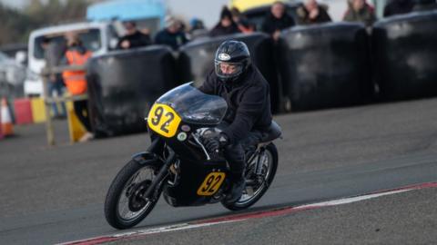 Ed Poole, a motorbike rider, he wears all blakc, and is on a black motorbike with the number 92 in a bright yellow bubble as he turns round a corner, there are spectators in the background.