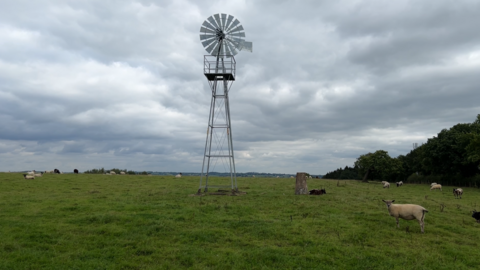 Windmill on a hill in a green field, with grey skies behind and sheep dotted about it