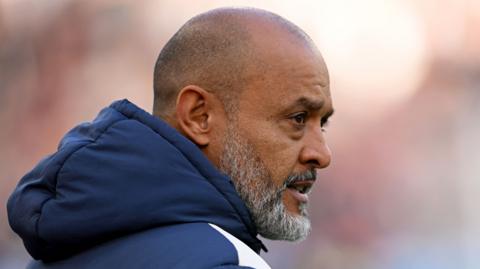A headshot of Nuno Espirto Santo with his face seen side on as he looks to the right. He is wearing a blue padded jacket with the large hood hanging behind his neck