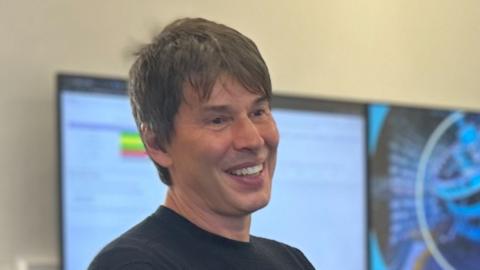 Brian Cox smiling in a room in Queen's University Belfast.  He has short dark hair and is wearing a black crew neck top 