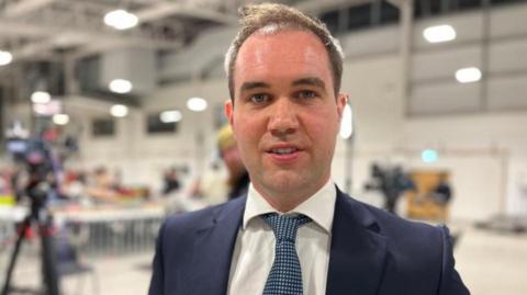 Oliver Lewis, who has short brown hair and is wearing a blue suit and tie looking at the camera at an election count.