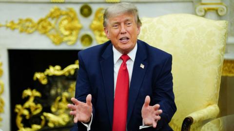 Donald Trump sits in the Oval Office wearing a navy blue suit and red tie.