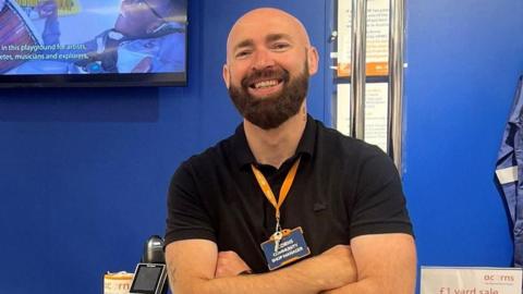A man with a shaved head and a lustrous dark beard stands in front of a shop counter with a white top and a blue frontage, labelled "acorns". He is wearing a black polo shirt and black jeans and has his arms crossed in front of his body. He is smiling at the camera.