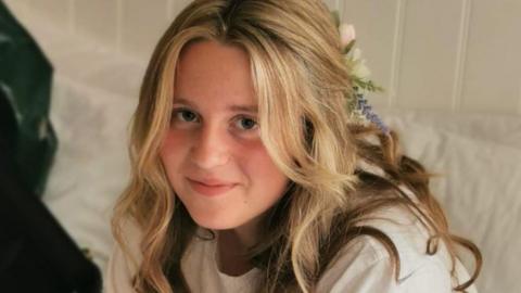 Chloe pictured wearing white t-shirt, and has a bow in her blonde hair. She is smiling at the camera