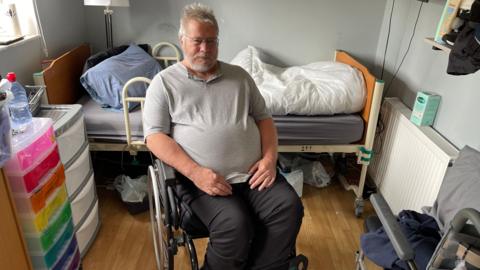 Paul Kitterman sits in a wheelchair in front of a hospital bed in his home. There are plastic drawers on one side of him and the bed has a blue pillow on it and a duvet. He is looking at the camera and wearing a grey t-shirt.