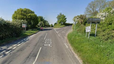 A shot looking down the A338 on a sunny day. A crossroads that lies diagonally. A road sign to the right points to Shalbourne. Roads are surroundd by lush greenery.