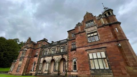 An imposing red sandstone building with stone archways and ornate features. There are decorative designs above the windows and a on the upper sections of the building's facade.