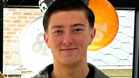 A young man smiling at the camera with short dark hair. He is standing in front of a window and there are balloons behind him.