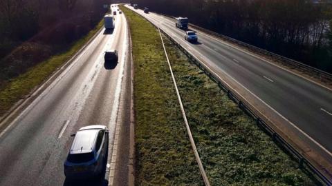 The A14 road near Ipswich: Two sections of  a back dual-carriageway. Traffic is travelling in both directions. The sun is glinting off the left-hand carriageway. A grass verge separates the two sections and grass and trees line either side of the road.