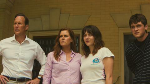 A man in a white shirt standing with his hands on his hps, a woman with long red hair and a pink shirt is standing his right. Ayoung woman with long brown hair in a white t-shirt is to her right. A young man with short brown hair is standing next to her. All four people are are standing in front of a house and looking into the distance.