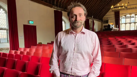 Tim FitzHigham stands in the performance space of Shakespeare's Guildhall in King's Lynn. Behind him are banks of red seats. Tim is wearing a pink shirt and has short brown hair and a beard. 