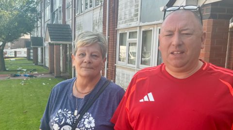 A man and a woman stand in front of a pile of rubbish on a front lawn by block of flats.