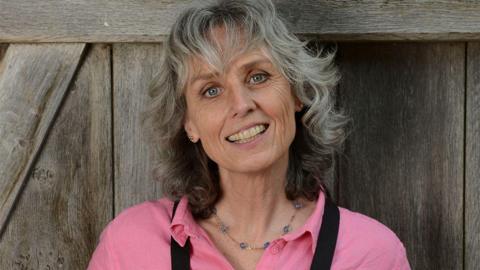 Suzie Fletcher pictured smiling against a wooden fence. She has grey wavy short hair, wears a pink shirt with a blue beaded necklace and a black apron.