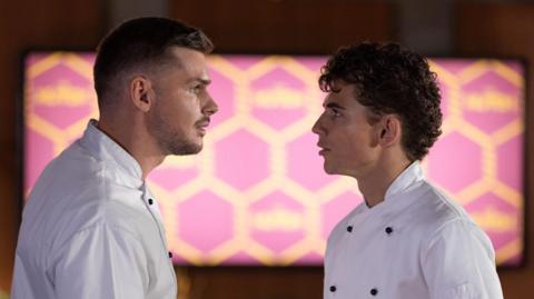 Two men with dark hair and white shirts looking at each other in front of an illuminated pink and yellow wall with a hexagonal pattern
