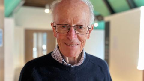 Close-up portrait of 90-year-old Hamish. He has short grey hair and glasses, smiling, wearing a dark sweater over a checkered shirt, standing indoors against a softly blurred background.