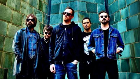 Andy Hall is on the extreme left of the picture alongside his band-mates in The Institutes. He has brown hair and a brown beard and is wearing sunglasses, as are four out the five people in the picture