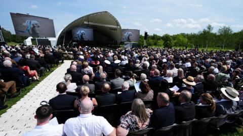 A large crowd of people are sitting in front of a stage which is covered with a tarpaulin canopy. There is a white walkway to the left hand side of the image. Behind the stage is a lawned area and a series of trees.