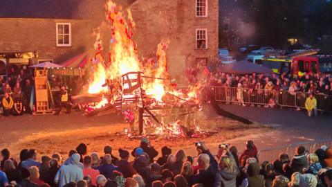 A wooden structure in the shape of a wolf which is covered in fire. It is standing in a village square on top of a pile of sand. Barricades form a circle around the fire, with crowds of people watching on. A fire engine can be seen parked in the background.