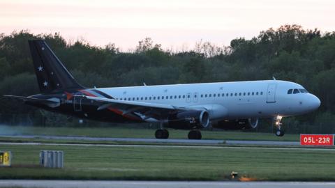 A plane with a white body and blue tail with white stars is seen from the side on a runway