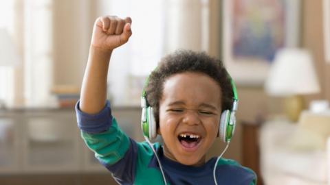 A boy in a navy blue and green jumper wearing green headphones with his eyes shut raises his arm listening to music.
