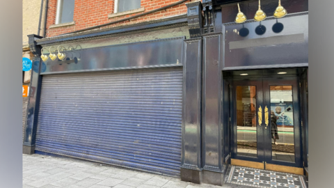 The entrance of a shop building with blue shutters down. To the right of it is a black framed door, with gold handles.