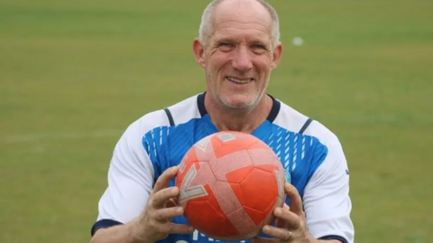 Roy holding a red football wearing a blue and white Posh jersey