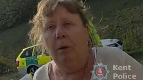 A woman is outside near a roadside, with a marked police vehicle behind her. She is looking at the camera. The image has a Kent Police logo on it.