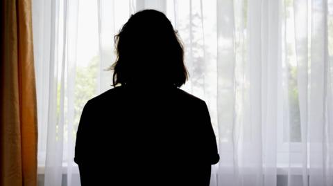 A stock image showing a silhouette of a woman with her back to the camera looking out of the window, which has net curtains.  