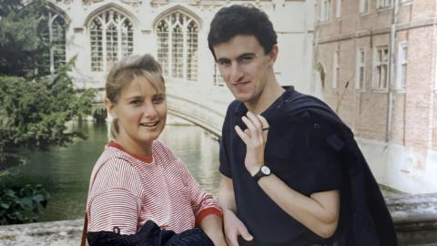 Rose Grant with her boyfriend Tim Burman. She is wearing a red and white stripey long-sleeved top. Tim is wearing a dark polo shirt and is holding a jacket over his left shoulder. They are stood on a bridge over a river or canal with old buildings behind them.