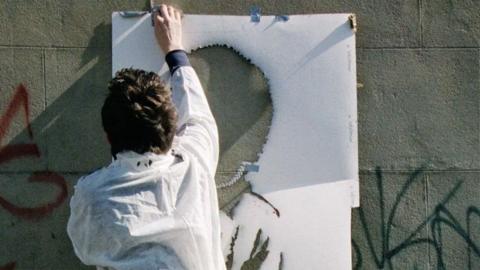 The back of a man wearing white overalls putting up a stencil on a wall