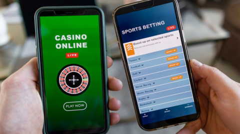 A close-up image of two mobile phones, each being held by a hand. The left mobile has a mainly green screen featuring the words casino online. The phone on the right has a predominantly blue screen including the words sports betting.