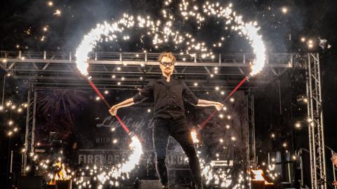 A person holding batons which have sparklers coming out the end of them. He is wearing all black and is stood on a stage at a fireworks display event.