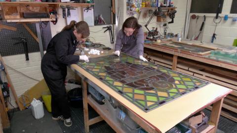 Two women working a piece of stained glass on a bench