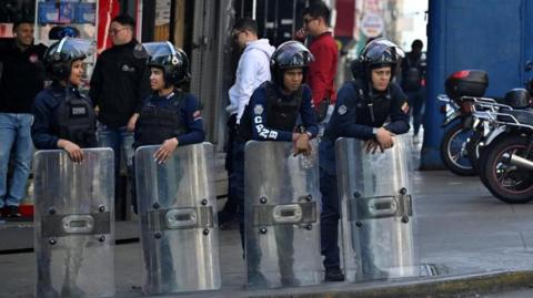 Police officers stand guard near the National Assembly in Venezuela's capital, Caracas. They are chatting to one another while wearing helmets and holding large shields.