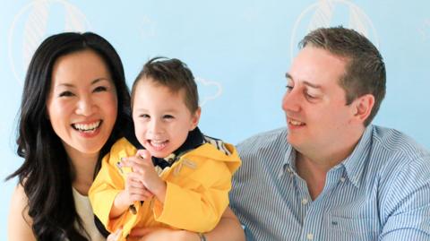 A family of three smiles during a photoshoot. There is a woman on the left holding a toddler wearing a yellow coat - they both look at the camera. The man on the right wears a striped shirt and looks towards the child.