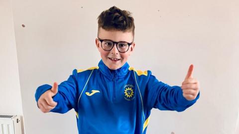 Leo Moseley stood with his thumbs up looking at the camera. He is wearing glasses and is smiling. He is wearing a blue and yellow tracksuit top.