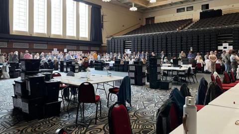 Wide view inside a large indoor hall set up for an election count, with rows of tables arranged on a patterned floor, ballot boxes stacked nearby and groups of people sorting and counting papers, with tiered seating and tall windows along one wall