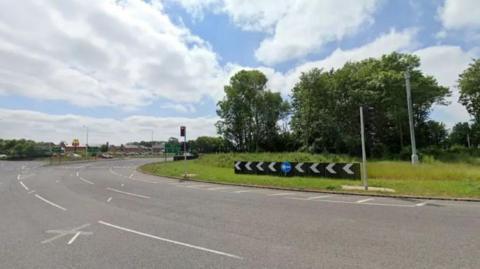 Google Streetview image of a three-lane roundabout, which has trees and grass in the middle.