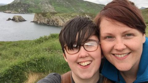 Lyra McKee and Sara Canning pose for a selfie. They are standing cheek to cheek, Lyra on the left, Sara on right. Lyra is wearing glasses and has short dark hair, sara is wearing a blue top and is smiling too. They are standing in front of cliffs, sand dunes, and the sea.