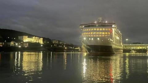 The Manxman, which is a large red, white, and black ferry, sitting in low water in Douglas Harbour at night. The lights from the ship, the harbourside buildings are reflecting on the water around it.