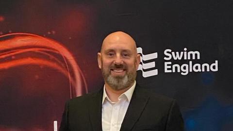 Ben wearing a black suit holding a wooden award standing in front of a red and black Swim England banner.