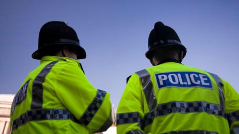 The backs of two police officers wearing hi-vis jackets and police helmets with blue sky in the background.