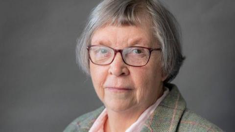 Councillor Jill Rhodes, a woman with grey hair wearing glasses and a tweed jacket. She is standing against a dark grey backdrop.
