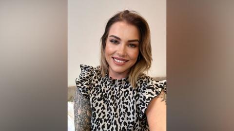 Woman with a large tattoo on her right arm smiling for the camera. She has brown hair. Her top has a leopard spot design.