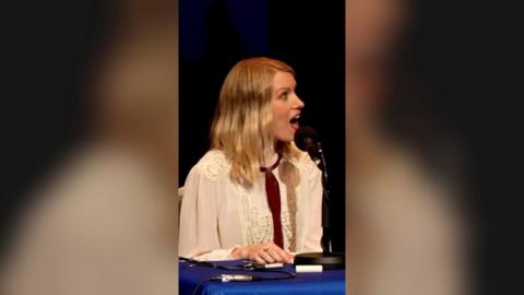 Rachel Parris, a woman with blonde hair and a white shirt, sits in front of mic, open mouthed in shock.