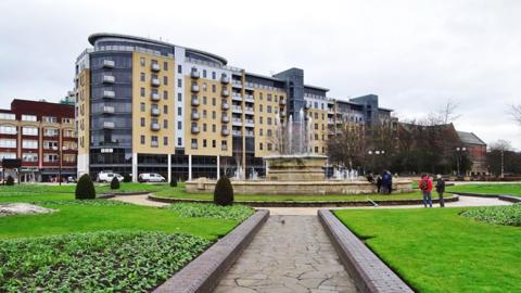 Queen's Court in Hull with yellow brickwork and grey steel cladding many of the flats have balconies and the penthouses are glass fronted. In the foreground is grass and a large fountain.
