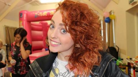 A young woman with red hair and blue eyes is smiling at the camera. She wears pink lipstick and is dressed in a white Mickey Mouse shirt and a black leather jacket. In the background there is a pink bouncy castle and it looks like a children's party is taking place.